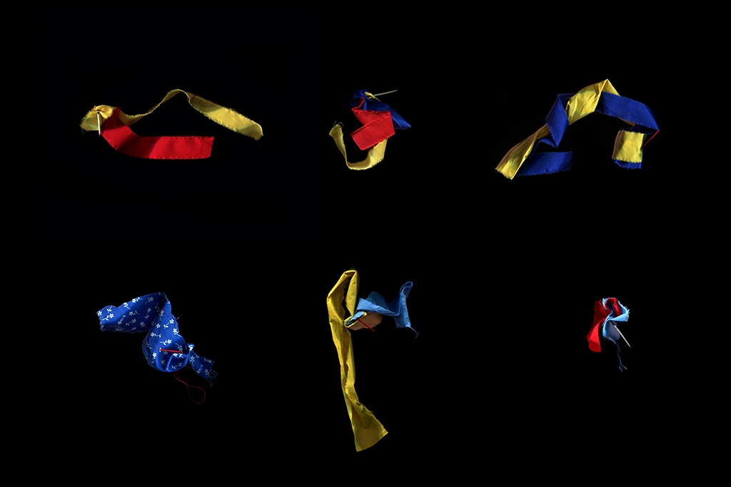 This image is composed of a closeup of six bundled sets of one-inch wide woven cloth strips of red, yellow, blue colours, positioned on a black surface. Each bundle of cloth is pierced by a porcupine quill that holds them together. The bundles are displayed equally spaced in two rows.