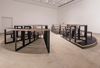 A view of the first configuration of furniture in Adrian Blackwell's Furnishing Positions series, from the side, facing into the gallery away from the entrance. The furniture has a minimalist design, with black metal legs, laminated wood surfaces (painted black on top), and open sides, making the structure's assembly visible from most angles.