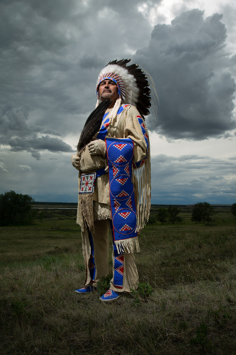 Stimson stands proudly, photographed from waist-level, in a Prairie landscape with stormy gray and blue clouds overhead. He wears Blackfoot dress with pale tanned hides and red, white, and blue beaded details, including a feathered headdress and jacket with long fringe. With hands clasped in front of his chest, he holds a large eagle feather and looks intently above the camera.
