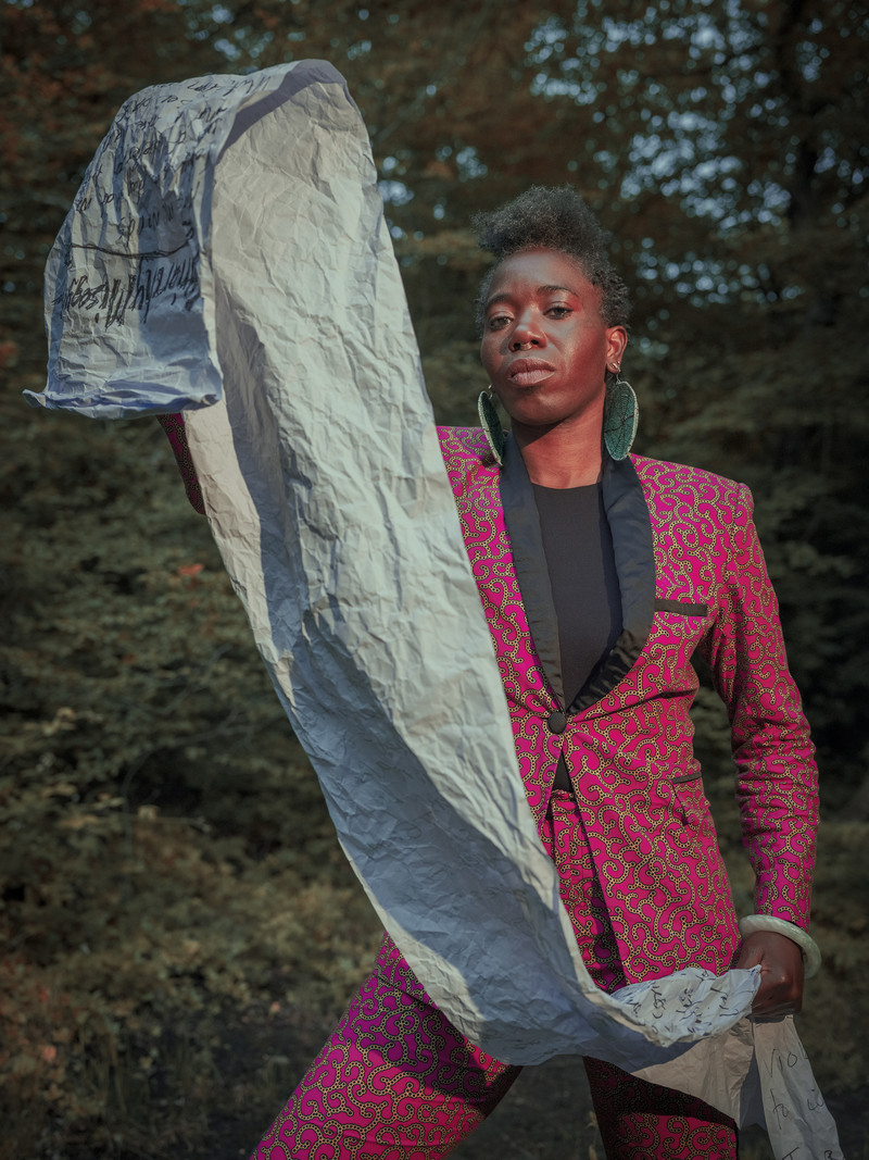 d’bi.young anitafrika stands looking defiantly at the camera, lips pursed, holding a long, deeply creased scroll that cascades from just above head height to their left, diagonally in front of hxr. d’bi wears a fuchsia suit with a yellow ornamental print, and large circular beaded earrings, standing in a forested area. The scroll has illegible handwritten text, and falls out of frame at the bottom right.