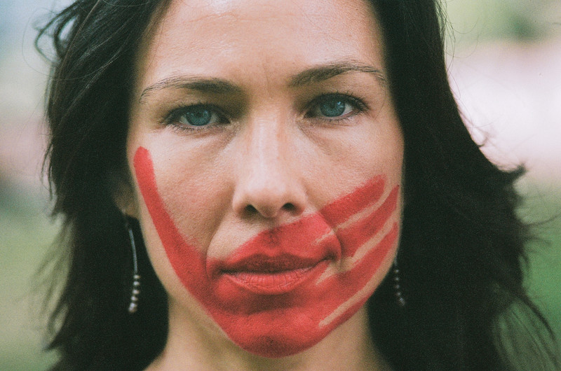 Emily Johnson’s face is seen close-up, with a red palm print painted over her mouth and jaw. The print is oriented diagonally, with the thumb nearly reaching her right eye. She looks into the camera intently with her mouth closed, and wears hoop earrings with beads. The image is soft focus and grainy.