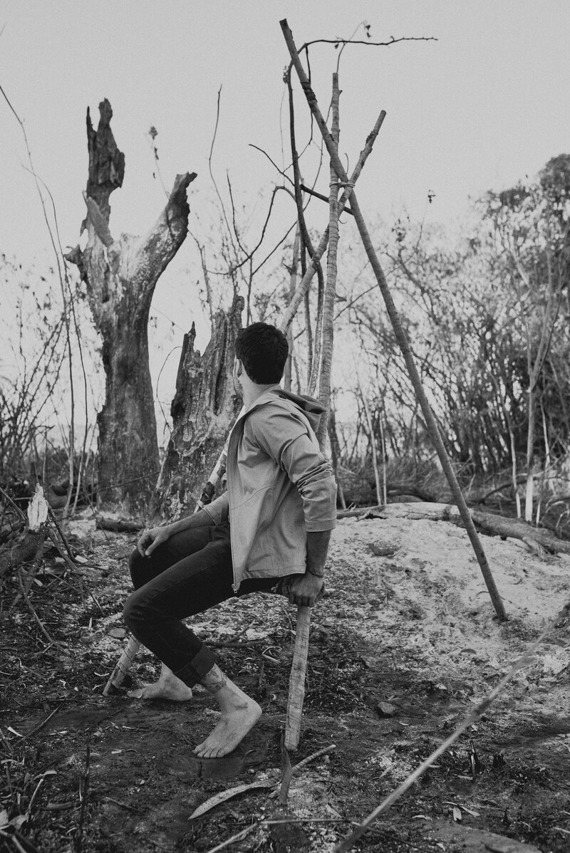 In a black-and-white image shot on film, Paulo Tavares is seen seated on a log structure, looking away from the camera. Barefoot and in jeans and a hooded jacket, Tavares is seen on a structure comprised of three vertical logs, set in a tripod formation with a crossbeam. The scene is overcast, in a a scrubby, perhaps recently burnt landscape.