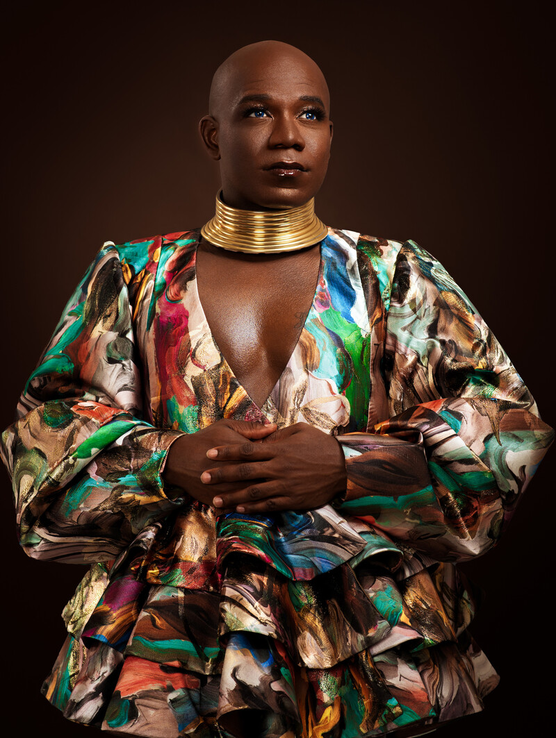 In a studio portrait with dark brown background, Ravyn Wngz stands poised with hands clasped in front of her chest, looking off to the right, above the camera. She wears an Ndebele-style gold choker necklace, and a low-cut shirt-dress with ruffles at the bottom and a colourful brushstroke print.