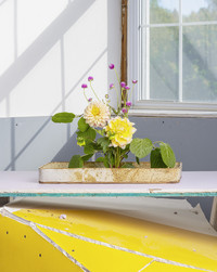 Yellow, white, and purple flowers emerge from an off-white piece of pottery in the centre of the image. The table underneath them is bright yellow and cracking. The window in the background casts a friendly shadow across the wall.