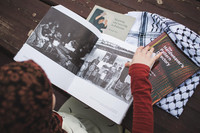 Photographed from above, a person sits at a wood table, looking at a book about Palestinian textiles. The book is accompanied by two other books entitled Spinning and Weaving in Palestine and The Handweaver’s Pattern Directory, as well as a Keffiyeh scarf.