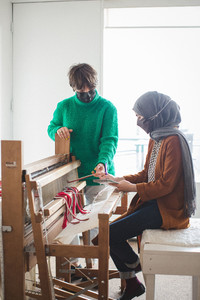 Two people are at a large loom. One sits in front of it writing in a notebook, wearing a grey hijab and brown sweater. The other person stands beside the loom, holding white and red threads coming from it.