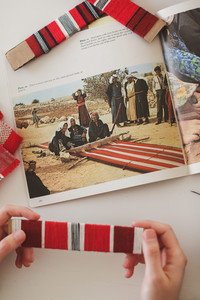 Photographed from above, a person holds a small cardboard warp beam with red, black, and white thread on it. Woven fabric and warp beams lay around the edges of the picture. In front of the person is an open book which shows an image of three Palestinian women weaving on a ground loom while a crowd of men and children smile around them.