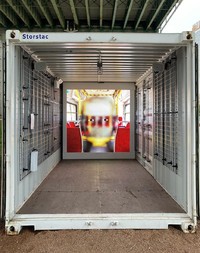 An image looking into a shipping container; on the end wall is a square projection of a face inside a public transit vehicle. The face has only cartoon-like eyes visible and is semi-transparent over the background, the result of being rendered in face-tracking software. 