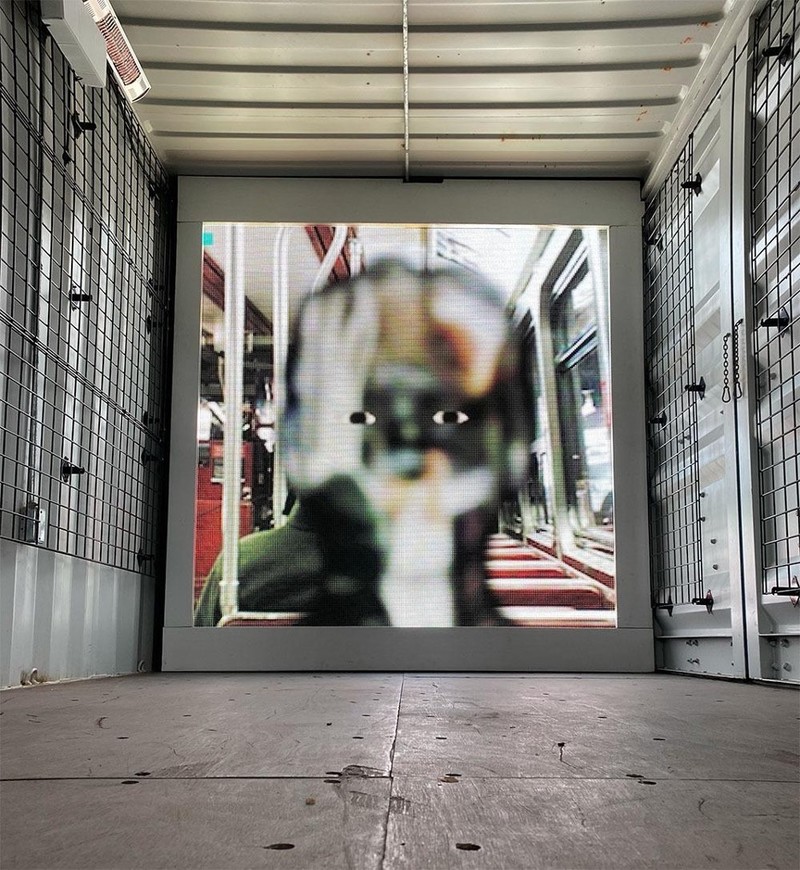 An image looking into a shipping container; on the end wall is a square projection of a face inside a public transit vehicle. The face has only cartoon-like eyes visible and is semi-transparent over the background, the result of being rendered in face-tracking software. 